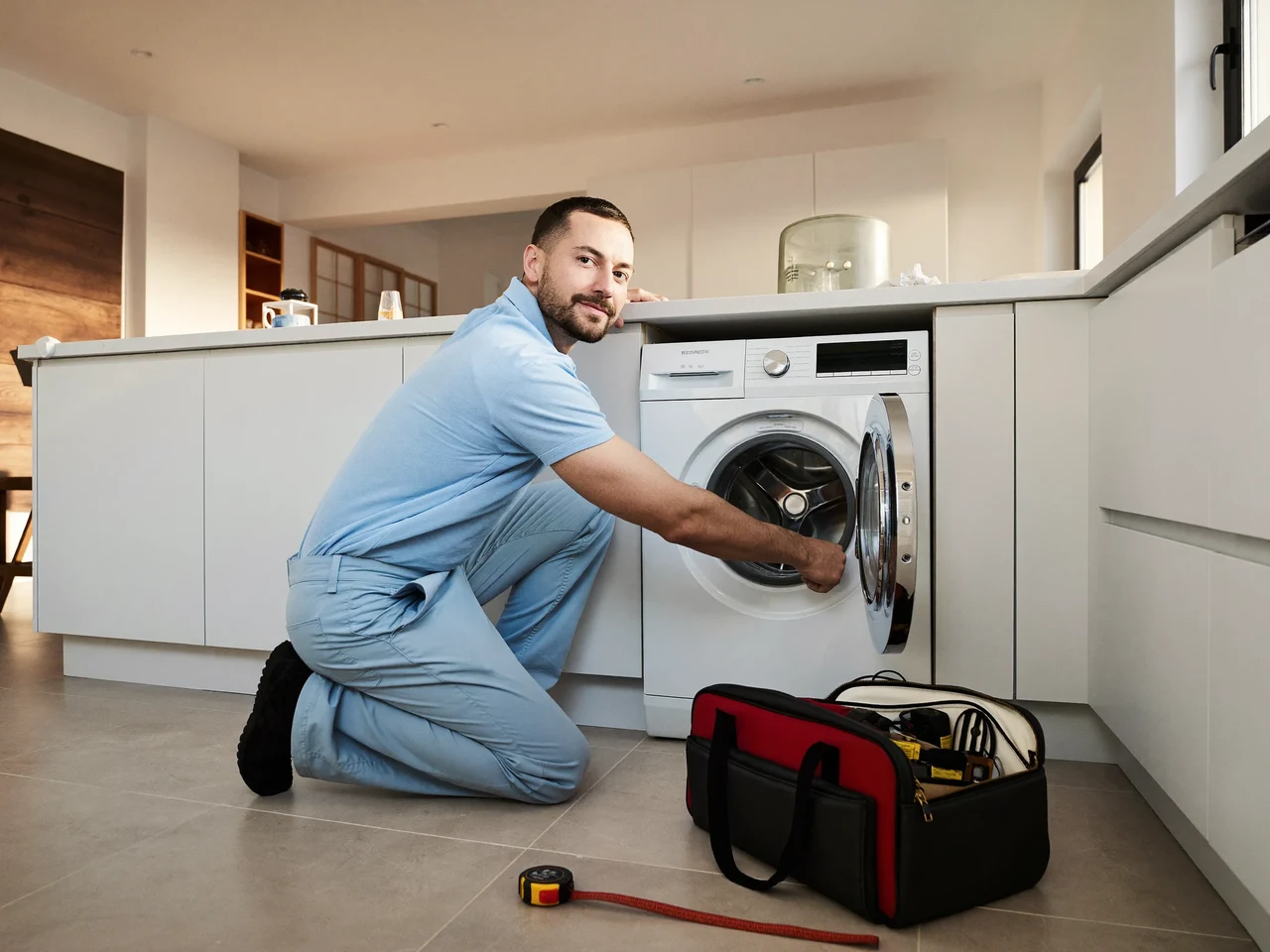 Tehnician în haine simple repară o mașină de spălat scoasă din nișă, într-o bucătărie luminoasă.